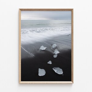 Op de afbeelding: Een ingelijste print met een zwart zandstrand en heldere ijsformaties. De oceaangolven zijn wazig, wat beweging suggereert. De lucht is bewolkt en de lijst is van licht hout.