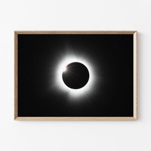 May include: A framed print of a solar eclipse. The image features a black disc surrounded by a bright white corona against a black background. The wooden frame adds a natural touch to the celestial artwork.