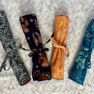 May include: Four fabric rolls with different patterns, tied with ribbon. The rolls are green with white swirls, blue with brown spots, orange with white spots, and blue with white leaves.
