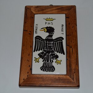 Op de afbeelding: Een ingelijste keramische tegel met een zwarte adelaar met een gele kroon en de tekst "PHS IMMOTAM MANET" in zwart.