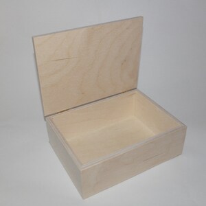 May include: A light-colored, unfinished wooden box with a hinged lid. The box is rectangular with a simple design, and the lid is partially open, revealing the interior. The wood grain is visible on both the box and the lid.