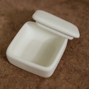 May include: A white ceramic box with a lid, the lid is open and the box is empty.