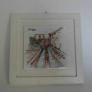 Op de afbeelding: Een witte ingelijste tegel met een aquarel-illustratie van een stenen gebouw met een waterpartij. De tegel is gelabeld met "L'Aquila".