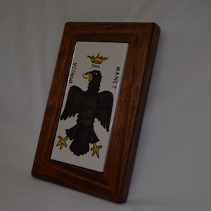 Op de afbeelding: Een ingelijste tegel met een zwarte adelaar met een gele kroon en de woorden "IMMOTTA MANET" in gele letters.