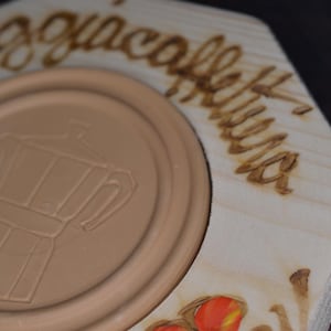 Könnte beinhalten: Ein Untersetzer aus Holz mit einem eingebrannten Design einer Kaffeekanne und den Worten "L'acqua e caffe'" in italienischer Sprache. Der Untersetzer hat eine braune Mitte mit einem erhöhten Rand und einen hellbraunen Außenring.