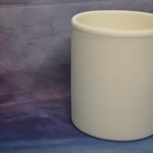Può includere: Un contenitore cilindrico in ceramica bianco sporco, con base arrotondata e bordo leggermente svasato. La superficie liscia e opaca contrasta con lo sfondo strutturato, multicolore viola e blu.
