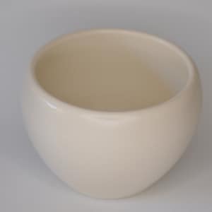 Può includere: Una ciotola in ceramica color crema a forma di uovo, con un esterno liscio e arrotondato. La ciotola ha un'ampia apertura e una superficie leggermente strutturata. L'interno è di una tonalità crema più chiara. La ciotola viene probabilmente utilizzata per servire o esporre.