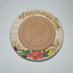 May include: A round wooden coaster with a brown ceramic insert. The coaster is decorated with a floral design and the text "Roaggia Caffeina". The ceramic insert has a silhouette of a coffee pot.
