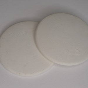 Może przedstawiać: Dwa białe ceramiczne podkładki pod kubki, jeden lekko zachodzący na drugi. Podkładki są okrągłe i mają gładką, lekko fakturowaną powierzchnię.