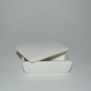 Puede incluir: Una pequeña caja rectangular blanquecina con una tapa ligeramente curvada, parcialmente abierta. La caja y la tapa tienen bordes redondeados y un acabado mate. La caja está vacía y se encuentra sobre una superficie blanca.