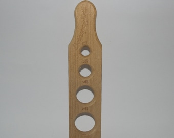May include: A wooden spaghetti measure with four differently sized holes. The handle is engraved with the words "Donna Spaghetti". The measure is made of light-colored wood and is designed to portion spaghetti.