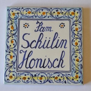 Può includere: Piastrella quadrata in ceramica con sfondo color crema e bordo decorativo. Il bordo presenta motivi floreali blu e gialli. La piastrella riporta le parole "Fam. Schiilin Honisch" scritte in blu.