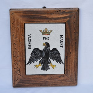 Op de afbeelding: Een ingelijst zwart-wit beeld van een zwarte adelaar met een gouden kroon erboven. De adelaar staat op een witte achtergrond met de woorden "IMMOTO" links, "PHS" in het midden en "MANET" rechts.