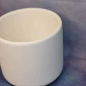 May include: A white, cylindrical ceramic mug with a smooth, matte finish. The mug has a rounded base and a wide opening. The background is a blurred mix of purple and blue hues. The mug is approximately 10 cm tall.