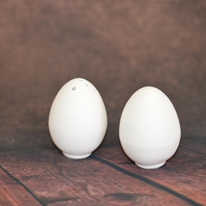 Peut inclure: Deux salières et poivrières blanches en forme d'œuf. L'une des salières a trois trous sur le dessus pour le poivre. Les salières sont sur une surface en bois avec un fond marron. Les salières sont en céramique.