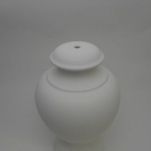 Könnte beinhalten: Weiße Keramikvase mit rundem Korpus und leicht ausgestelltem oberen Rand. Die Vase hat eine glatte, matte Oberfläche und ein kleines Loch oben.