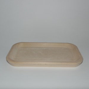 Può includere: Un vassoio rettangolare in legno con angoli arrotondati. Il vassoio è realizzato in legno chiaro e presenta una superficie interna scanalata. Il design è semplice e minimalista, adatto per servire o esporre.