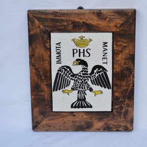 Op de afbeelding: Een ingelijste tegel met een zwart-witte adelaar met een gele kroon erboven. De tegel heeft de woorden "Immorta" en "Manet" aan weerszijden van de adelaar en de letters "PHS" onder de kroon.