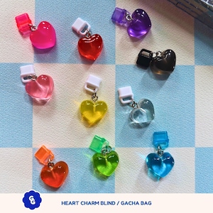May include: A set of ten heart-shaped charms in various colors, including red, pink, purple, black, yellow, green, blue, and orange. Each charm is attached to a small clip, making them perfect for adding to bags, backpacks, or other accessories. The charms are arranged on a checkered blue and white background. The text "HEART CHARM BLIND / GACHA BAG" is printed below the charms.