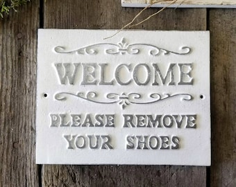 May include: White sign with black lettering that reads "Welcome Please Remove Your Shoes". The sign has a decorative border and is mounted on a wooden surface.