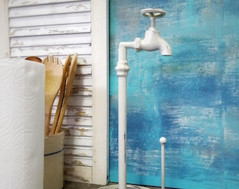May include: A white, vintage-style paper towel holder in the shape of a tap. The holder features a round base with decorative scrollwork and a vertical post. The tap handle is at the top. A roll of paper towels is visible to the left.