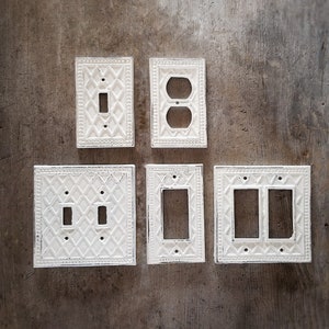 Könnte beinhalten: Fünf weiße Porzellan-Lichtschalter- und Steckdosenabdeckungen mit einem dekorativen Diamantenmuster. Die Abdeckungen sind in einer Reihe auf einer braunen Holzoberfläche angeordnet.