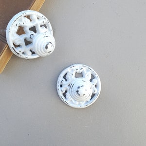 May include: Two white cast iron drawer pulls with a decorative design. The pulls have a distressed white finish and a round shape.