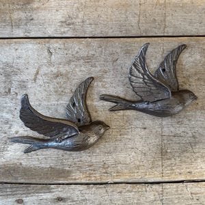 May include: Two cast iron bird sculptures in flight, with detailed wing and tail feathers. The birds are a dark, metallic gray color and are mounted on a weathered wooden surface. Decorative home decor.