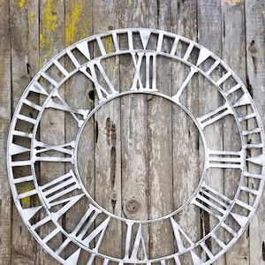 May include: A large, silver-colored metal clock face with Roman numerals. The clock face is hanging on a weathered wooden wall.