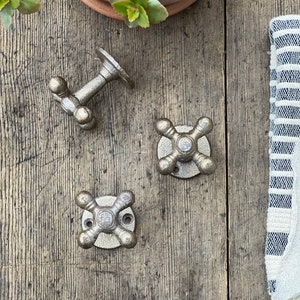 Peut inclure: Trois crochets en métal de style antique en forme de poignée de robinet vintage. Les crochets sont de couleur argentée et ont une finition vieillie. Ils sont montés sur une surface en bois.