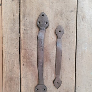 May include: Two rustic black metal door handles with pointed ends and decorative holes. The handles are mounted on a wooden surface.