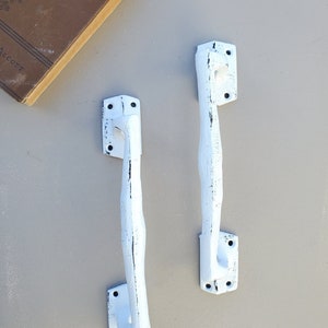 Könnte beinhalten: Zwei weiße Gusseisen-Türgriffe mit einer abgenutzten Oberfläche. Die Griffe haben ein schlichtes, elegantes Design und sind perfekt, um jedem Raum einen Hauch von Landhausstil zu verleihen.