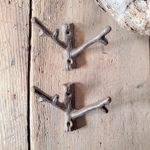 May include: Two rustic cast iron hooks shaped like tree branches. The hooks are mounted on a wooden wall.