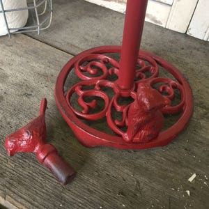 May include: A red cast iron paper towel holder with a decorative base featuring a squirrel and scrollwork. The holder is on a wooden surface. A separate red bird-shaped piece is next to the base.