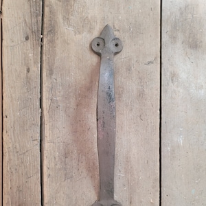May include: A black metal door handle with a fleur-de-lis design at the top and bottom. The handle is mounted on a wooden door.