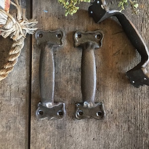 May include: Three rustic cast iron door handles with a dark brown finish. The handles have a simple, elegant design with a curved shape and a flat surface. The handles are shown on a wooden surface.