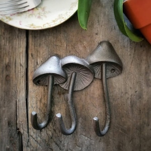 May include: A wall-mounted metal hook rack featuring three mushroom-shaped hooks. Each hook is cast in a dark gray finish with a textured cap and a curved stem that serves as the hook. The design displays detailed gills underneath the mushroom caps.