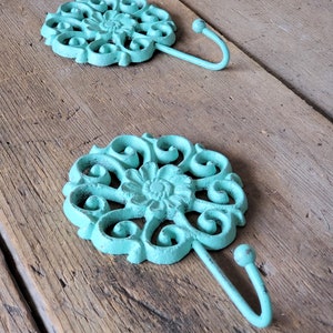 Puede incluir: Dos ganchos de metal azul turquesa con un dise&ntilde;o floral decorativo. Los ganchos est&aacute;n montados en una superficie de madera.