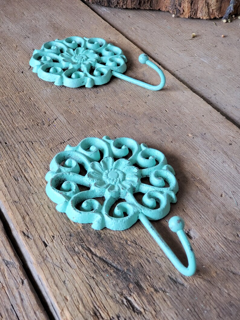 Puede incluir: Dos ganchos de metal azul turquesa con un dise&ntilde;o floral decorativo. Los ganchos est&aacute;n montados en una superficie de madera.