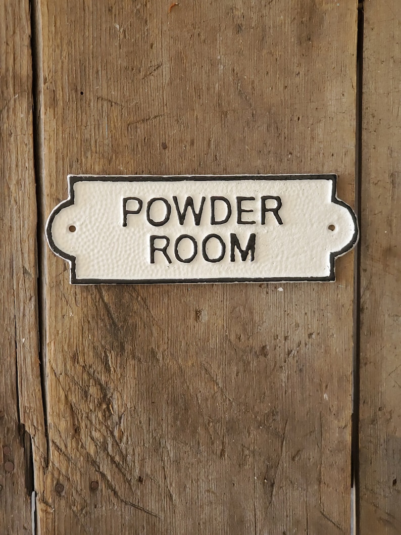 May include: A rectangular, off-white sign with the words "POWDER ROOM" in black lettering. The sign has a textured surface and a black border. It is mounted on a weathered, wooden door.