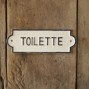 May include: A rectangular, off-white sign with the word "TOILETTE" in black lettering. The sign has a textured surface and a black border. It is mounted on a weathered, wooden background.