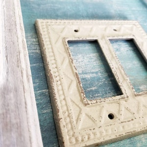 Op de afbeelding: Een witte, decoratieve, dubbele lichtschakelaar met een verweerde afwerking en een sierlijk ontwerp. De afdekplaat is gemonteerd op een lichtblauw houten oppervlak.
