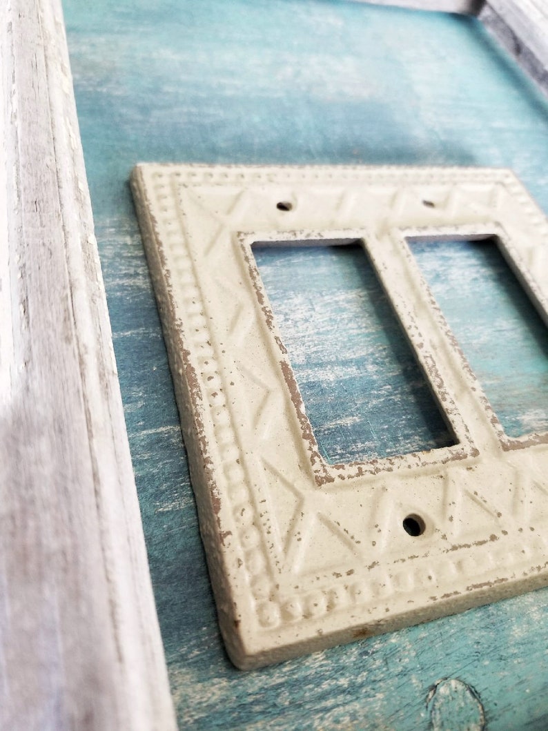Op de afbeelding: Een witte, decoratieve, dubbele lichtschakelaar met een verweerde afwerking en een sierlijk ontwerp. De afdekplaat is gemonteerd op een lichtblauw houten oppervlak.