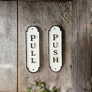 Op de afbeelding: Twee witte rechthoekige borden met zwarte randen en afgeronde hoeken. De borden zijn gemonteerd op een houten oppervlak. Het linker bord zegt "PULL" in zwarte letters. Het rechter bord zegt "PUSH" in zwarte letters.