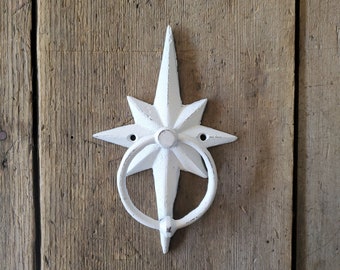 Puede incluir: Un llamador de puerta de hierro fundido blanco en forma de estrella con un anillo de tracción. El llamador está montado en una puerta de madera.