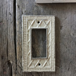 Op de afbeelding: Een witte, decoratieve lichtschakelaar-afdekplaat met een ruitpatroon en een parelrand. De afdekplaat is gemonteerd op een verweerd houten oppervlak.