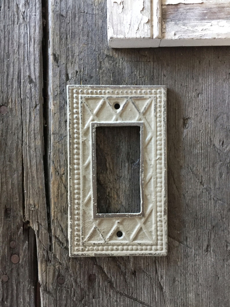 Op de afbeelding: Een witte, decoratieve lichtschakelaar-afdekplaat met een ruitpatroon en een parelrand. De afdekplaat is gemonteerd op een verweerd houten oppervlak.