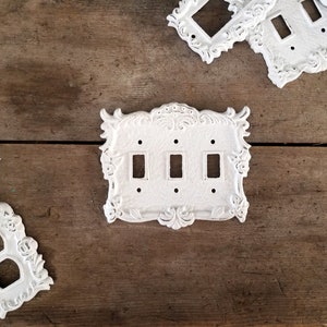 Puede incluir: Tres tapas de interruptor de luz de porcelana blanca con bordes decorativos ornamentados. Las tapas tienen tres y dos interruptores de palanca.