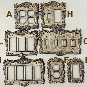 Könnte beinhalten: Acht dekorative Wandplattenabdeckungen in Silber mit verzierten Details. Die Platten sind mit A, H, L, M, O, F und E beschriftet.