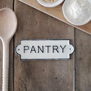 May include: A rustic pantry sign with the word "PANTRY" in black lettering. The sign is white with a black border and is mounted on a wooden surface. A wooden spoon, two small white bowls with oats and flour, and a wooden cutting board are also visible.